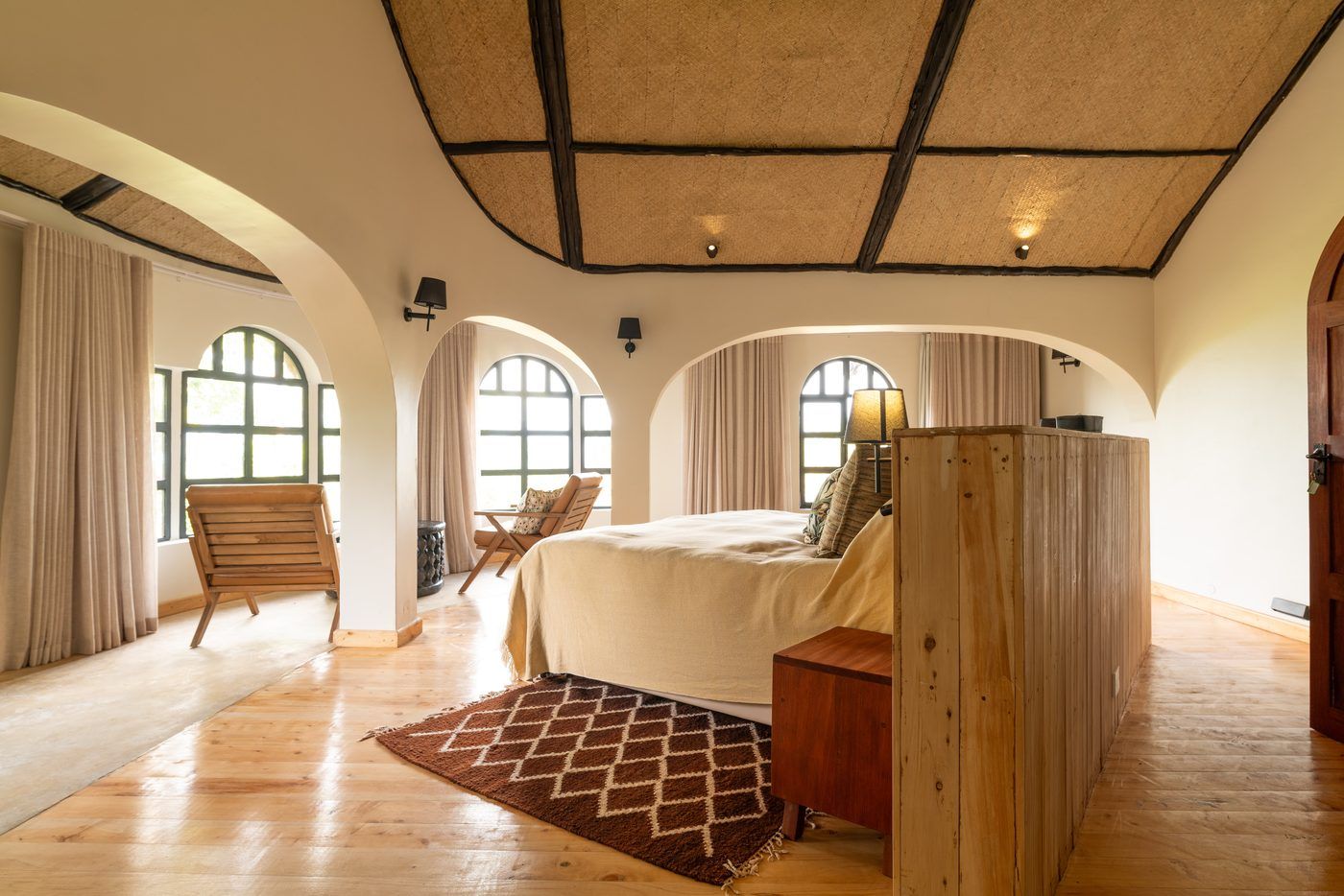 The upstairs bedroom at O'Leleshwa in daylight: three arched windows, a timber headboard partition and woven ceiling panels.