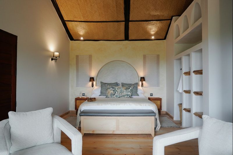 An O'Lerai bedroom — arched plaster headboard wall with twin sconces, a king bed with patterned cushions, cream armchairs in the foreground.