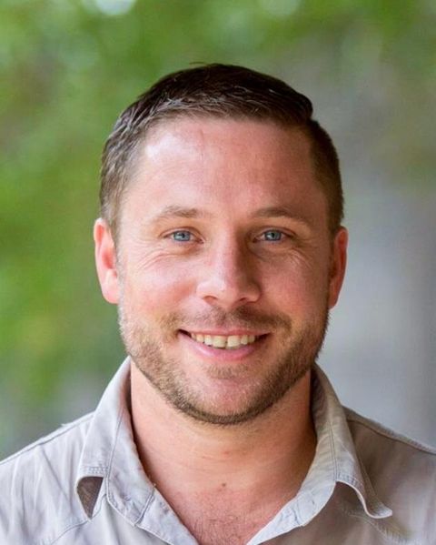 Neil Steedman smiling in a khaki shirt, green foliage soft behind him.