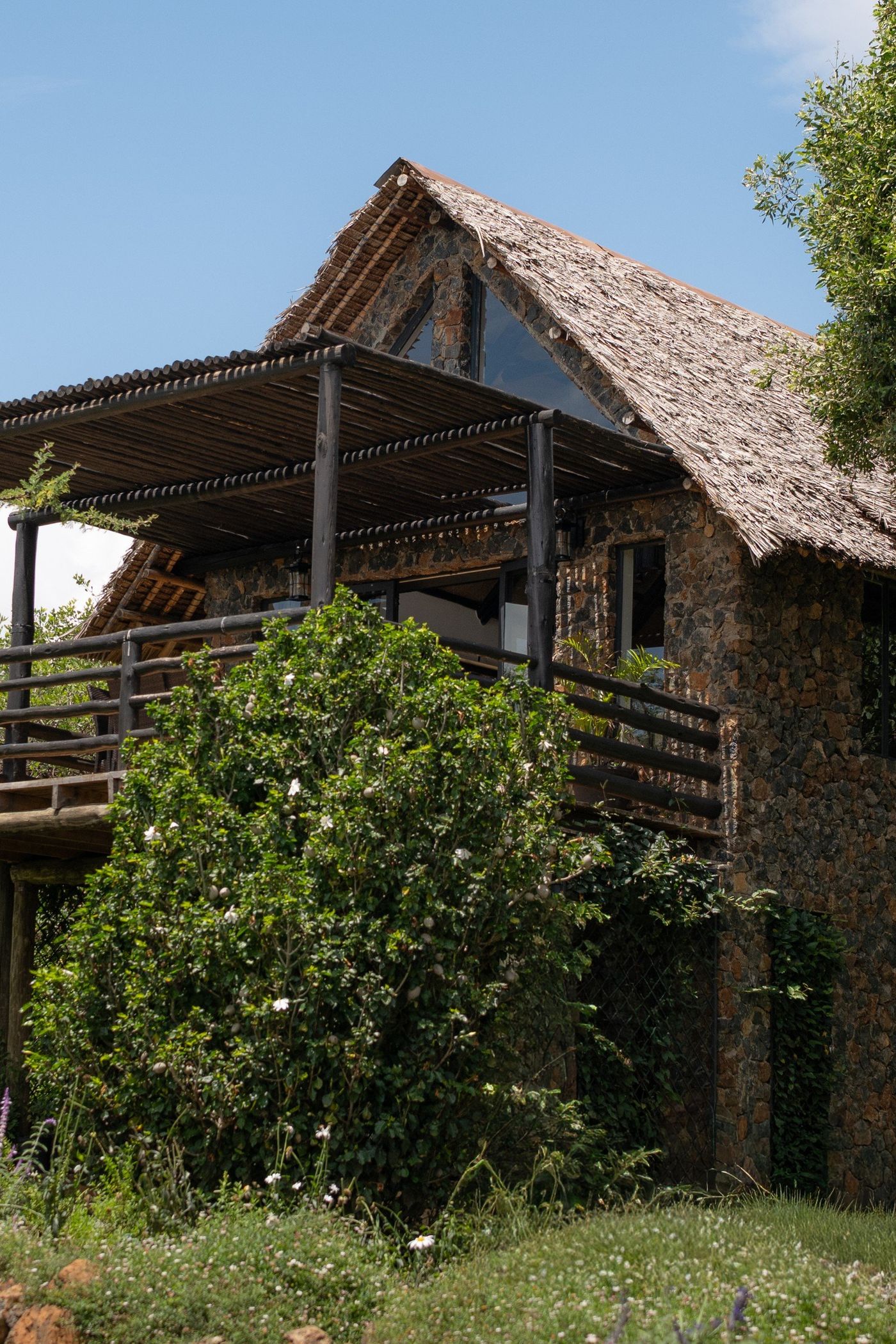 O'Lorien from the lower garden — rough estate stone under the thatched gable, the slatted pole pergola over the deck, white roses on the balustrade.