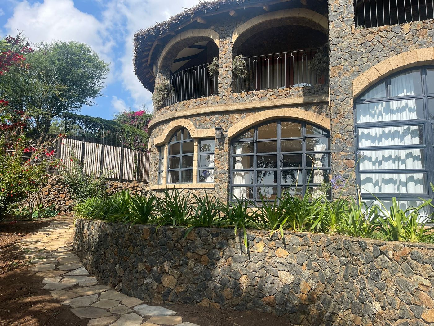 The stone face of O'Leleshwa in morning light — arched crittall windows, the curved balcony above, agapanthus in the raised bed along the path.
