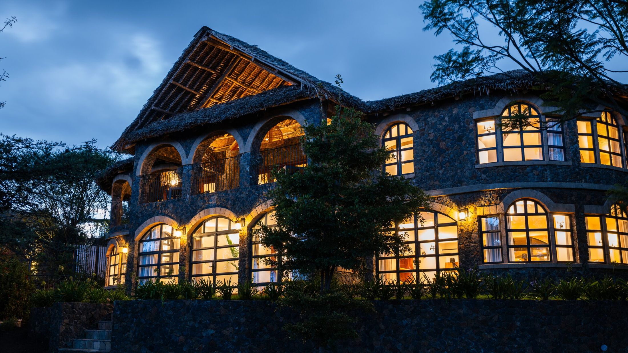 O'Leleshwa villa at nightfall — stone arches under makuti thatch, every arched window lit warm against a deep blue sky.