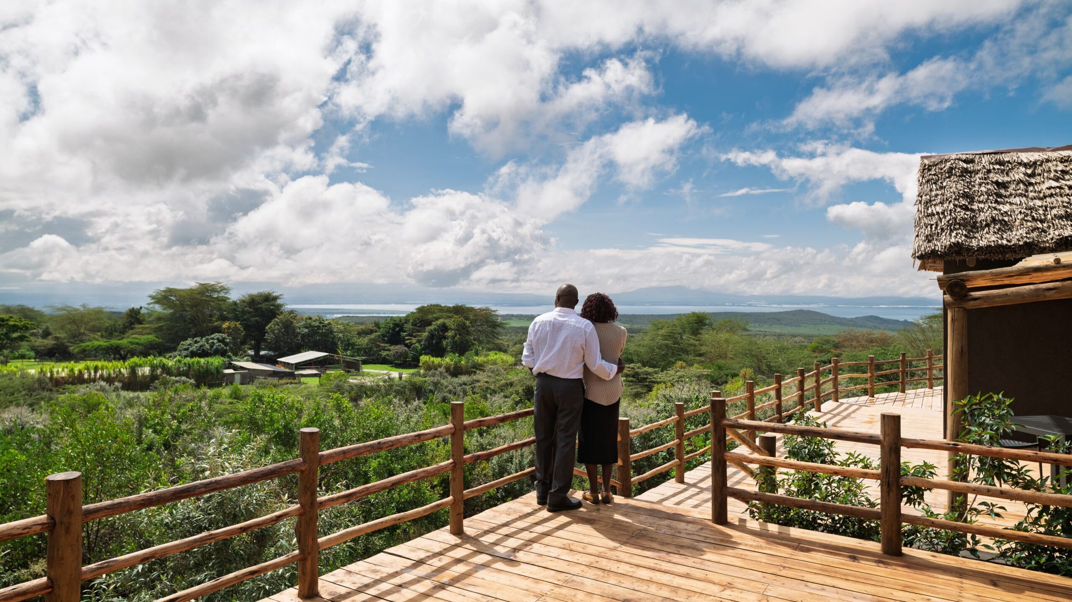 Two people at the timber deck rail at O'Lerai, looking over the bush canopy to Lake Naivasha under cumulus clouds.