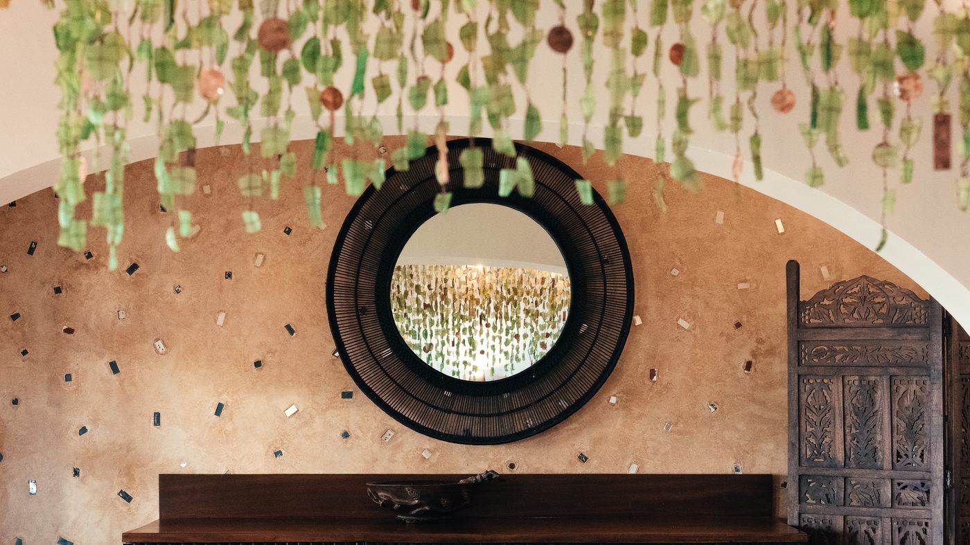 Strands of repurposed Malawian glass hanging past a round woven black mirror on mirror-flecked plaster.