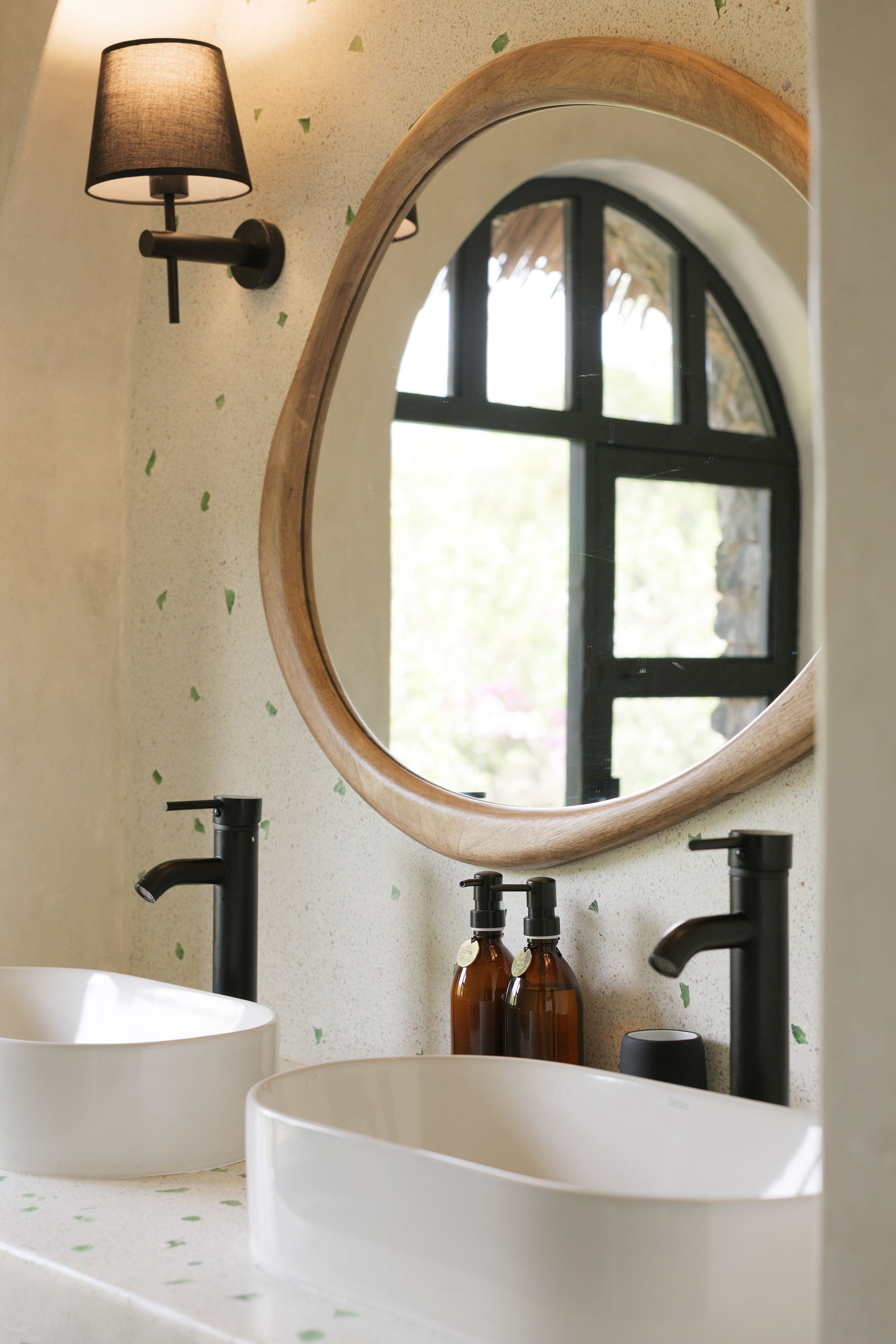 Twin white basins with black taps on a terrazzo counter flecked with green glass, an organic wood-framed mirror reflecting the arched window and thatch.