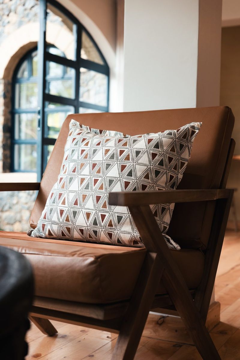 A tan leather armchair with a triangle block-printed cushion beside a blue steel-framed arched window, stone wall beyond.