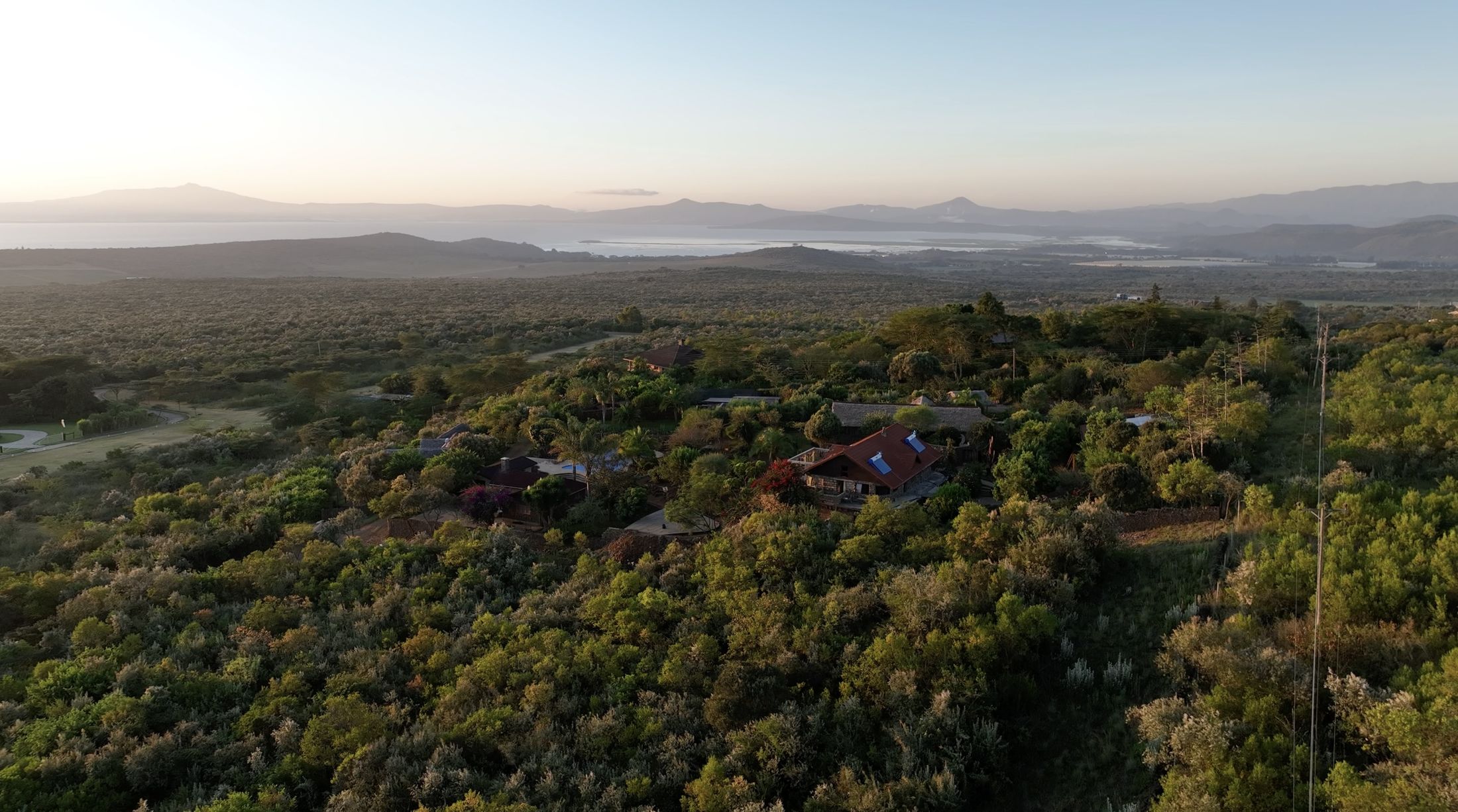 Dawn aerial of the hilltop estate in silver-green leleshwa scrub, Lake Naivasha and the Rift hills pale beyond.